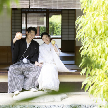 茶室の縁側に並んで座り、手を挙げて笑う紋付袴の新郎と白無垢の花嫁。緑に包まれた白鳥庭園の和装フォトウェディング
