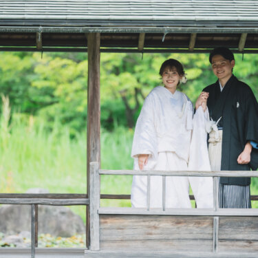 屋根付きの渡り廊下で、新郎の袖に手を添えて並ぶ白無垢の花嫁。緑の庭を背にした白鳥庭園でのフォトウェディング