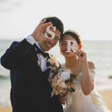 海を背にイニシャルのオブジェを顔の横に掲げ、笑顔を見せる新郎新婦。ビーチ前撮りで人気の小物使い