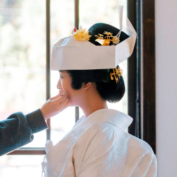 ヘアメイクスタッフが綿帽子姿の花嫁に紅を差している支度風景。鶴の刺繍が入った白無垢と、金色のかんざしのクローズアップ。