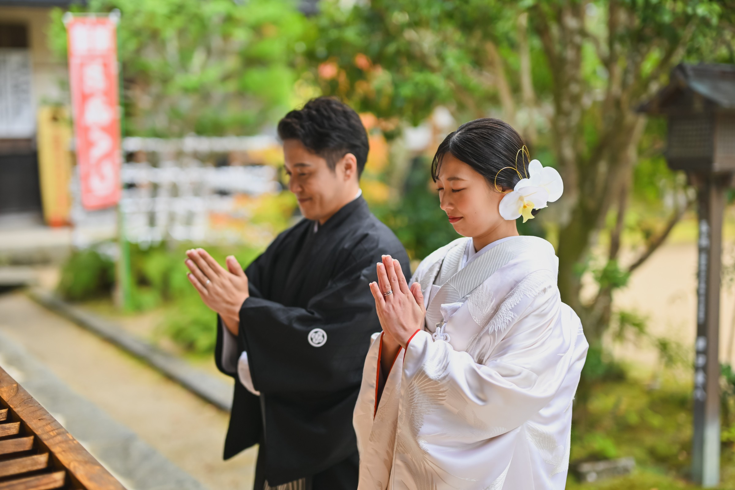 神社の境内で目を閉じ、静かに手を合わせてお参りする黒紋付袴姿の新郎と白無垢姿の花嫁。指輪をはめた手元が印象的な一枚。