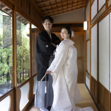 欄間の透かしが美しい畳の廊下で、扇子を持つ新郎と白無垢の花嫁が寄り添う。白鳥庭園の茶室で撮る和装前撮り