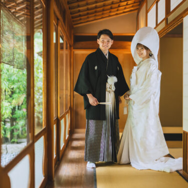 すだれ越しに緑が見える畳の廊下で、綿帽子に白無垢の花嫁と紋付袴の新郎が並ぶ和装前撮り。白鳥庭園の和室で撮る結婚写真