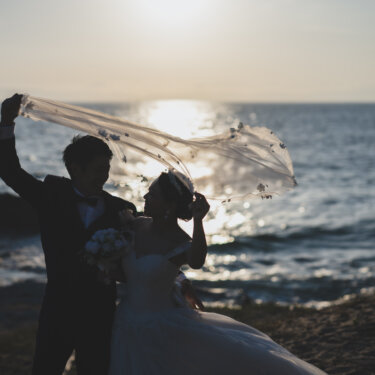 夕日の海を背に、新郎が花嫁のロングベールを広げる逆光のシルエット。知多半島の海で撮るドラマチックな前撮り