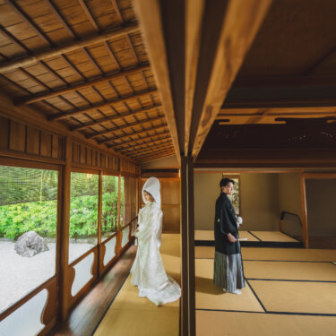 石庭に面した長い縁側に、綿帽子と白無垢の花嫁、離れて立つ紋付袴の新郎。奥行きを生かした和室での和装前撮り