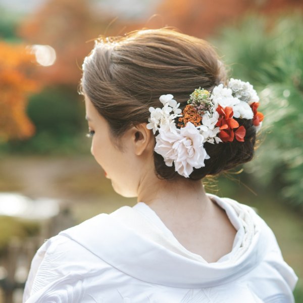 白とオレンジの花をあしらったまとめ髪。紅葉を背景にした白無垢姿の花嫁の後ろ姿クローズアップ。
