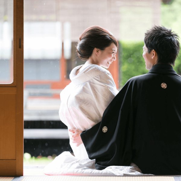 畳の部屋で正座し、笑顔で見つめ合う黒紋付袴姿の新郎と白無垢姿の花嫁の後ろ姿。障子越しに朱色の欄干が見える和の風情ある一枚。