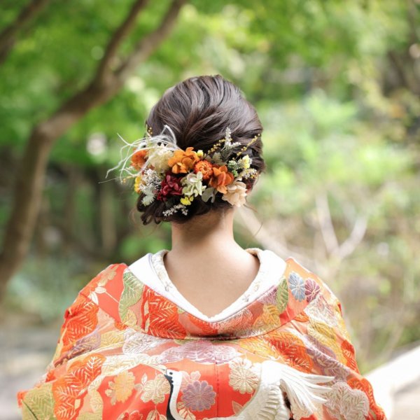 オレンジ系の小花をあしらったまとめ髪。鶴の刺繍が入った朱色の色打掛姿の花嫁の後ろ姿クローズアップ、緑豊かな並木道を背景に。