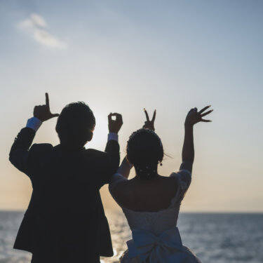 海に向かって手で'LOVE'の文字を作る新郎新婦の後ろ姿のシルエット。遊び心のあるビーチのウェディングフォト