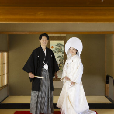 掛け軸のある床の間の前の赤い毛せんに並ぶ、扇子を持つ紋付袴の新郎と綿帽子の白無垢の花嫁の和装結婚写真