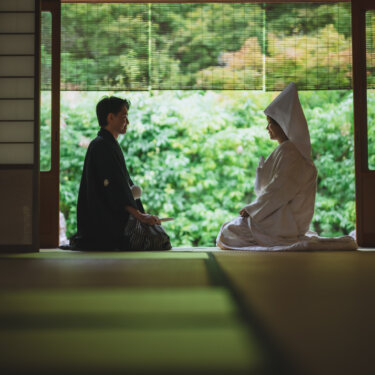庭の緑を背に畳の上で向き合って座る、綿帽子の花嫁と紋付袴の新郎のシルエット。白鳥庭園の和室で撮る和装の結婚写真