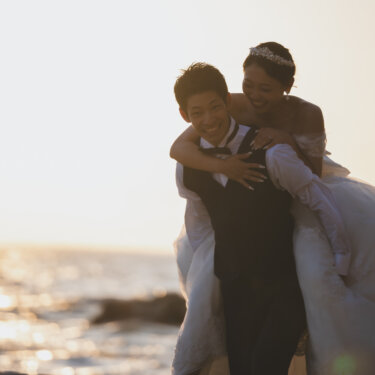 夕日に照らされた海辺で、新郎の背中に飛び乗っておんぶされる花嫁。笑顔あふれる遊び心のあるビーチ前撮り