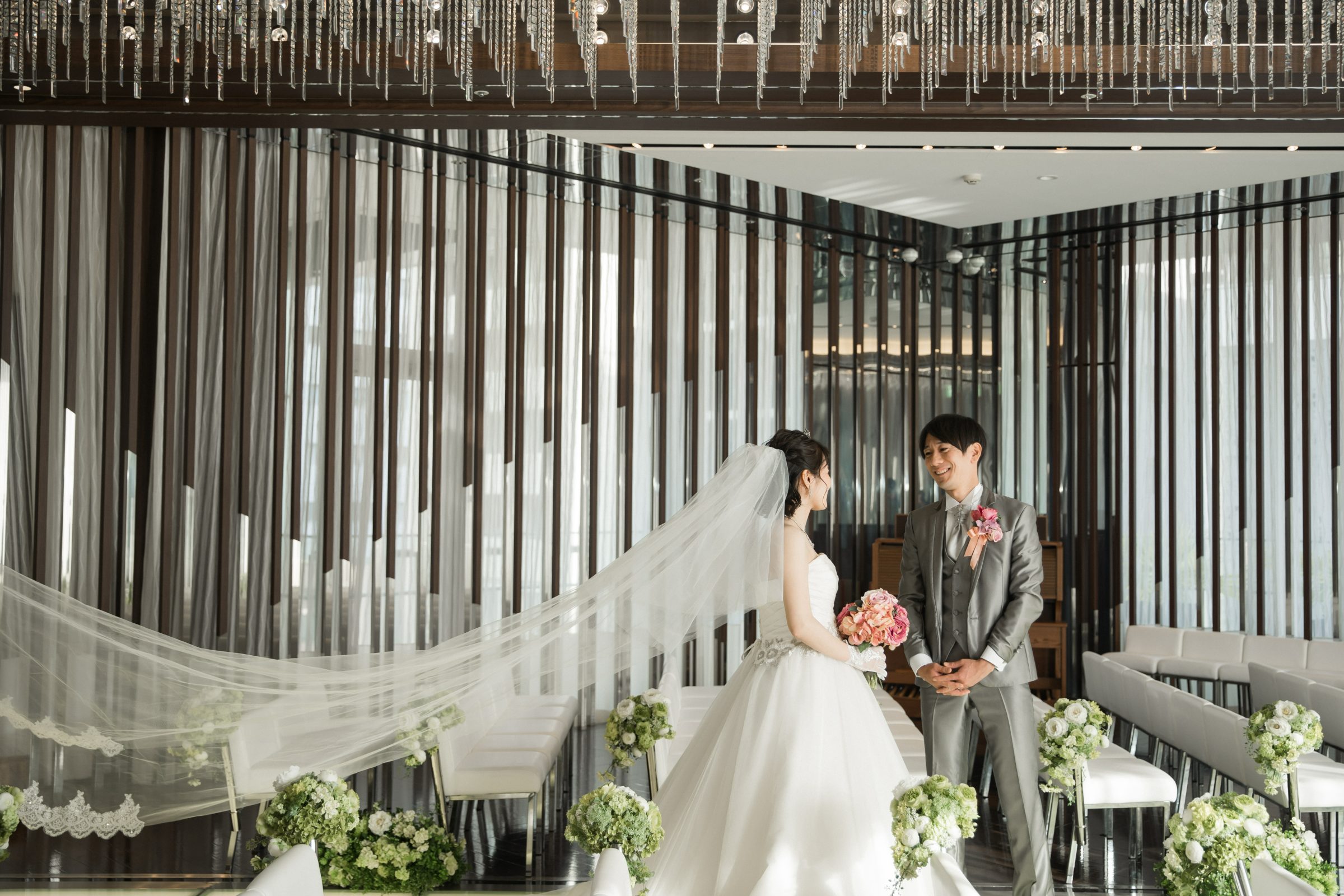 シャンデリアが輝くチャペル内で、ピンクのバラのブーケを持つ新婦とグレーの三つ揃いスーツの新郎が見つめ合う、長いベールがなびくチャペルフォトウェディング。