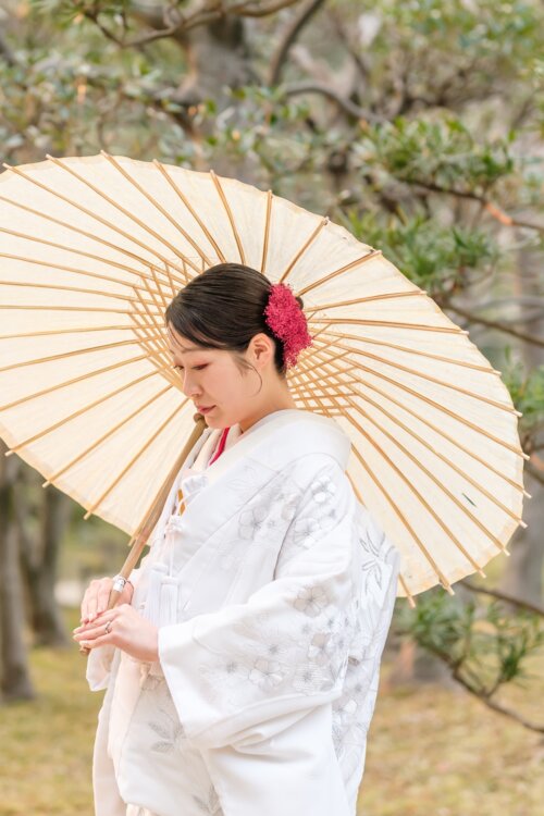 日本庭園の木々の下で和傘を差す、赤い髪飾りを合わせた白無垢の花嫁。ロケーションフォトで人気の和傘ポーズ