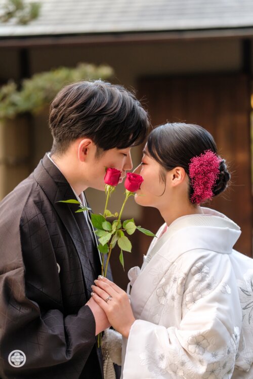 赤いバラを手に寄り添う黒紋付の新郎と白無垢の花嫁。花の香りを楽しむ自然な表情の和装前撮り