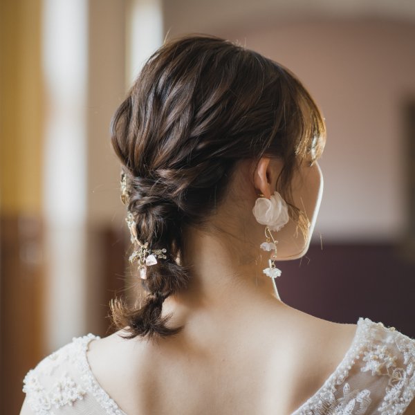 編み込みでまとめたルーズなヘアスタイルと、白い花モチーフの揺れるイヤリング。ドレス姿の花嫁の後ろ姿クローズアップ。