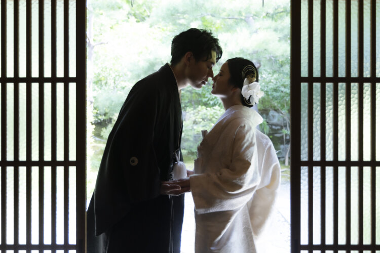 格子戸の間から、額を寄せ合う紋付袴の新郎と白無垢の花嫁のシルエット。白鳥庭園の緑が映える和装前撮り