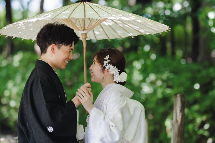 白い和傘の下で手を取り合い見つめ合う、黒紋付袴の新郎と白無垢の花嫁。白鳥庭園の緑に包まれた和装前撮り