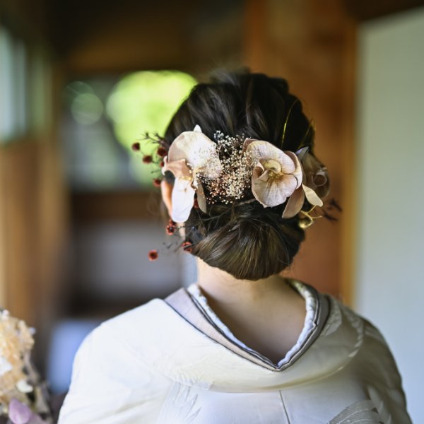 くすみカラーの大ぶりな花飾りをあしらったシニヨンヘア。白無垢姿の花嫁の後ろ姿クローズアップ。