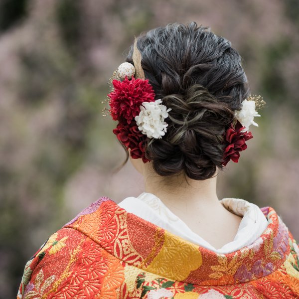 赤と白の椿の花飾りをあしらった編み込みまとめ髪。朱色の色打掛姿の花嫁の後ろ姿クローズアップ。